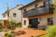 Ein modernes Einfamilienhaus mit einem markanten Balkon aus Holz. Der Eingangsbereich ist mit Terrakottaplätten gestaltet, umgeben von gepflegten Sträuchern und Blumen. Im Vordergrund steht ein Bohrgerät, das auf den Garten zeigt und auf mögliche Renovierungsarbeiten hinweist.