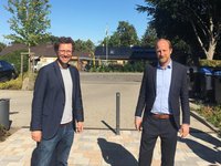Zwei Männer stehen auf einem gepflasterten Weg. Der linke Mann trägt eine Brille und ein gestreiftes T-Shirt unter einem Blazer, während der rechte Mann in einem hellblauen Hemd und einem dunklen Anzug gekleidet ist. Im Hintergrund sind Bäume und Gebäude zu erkennen.