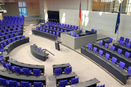 Bildbeschreibung: Der Innenraum eines Parlaments mit mehreren Sitzreihen in lila und grauen Farben. Im Vordergrund befindet sich ein Bereich mit einem Rednerpult. Im Hintergrund stehen Flaggen Deutschlands und der EU. Der Raum ist modern und gut beleuchtet.