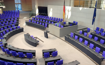 Bildbeschreibung: Der Innenraum eines Parlaments mit mehreren Sitzreihen in lila und grauen Farben. Im Vordergrund befindet sich ein Bereich mit einem Rednerpult. Im Hintergrund stehen Flaggen Deutschlands und der EU. Der Raum ist modern und gut beleuchtet.