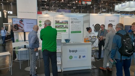 Messestand mit mehreren Personen, die sich um einen Tisch versammeln. Ein Bildschirm zeigt Informationen, während Informationen auf Plakaten an der Wand präsentiert werden. Der Stand gehört zur Firma bwp und bietet Dienstleistungen an.