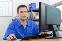 Ein Mann in blauer Arbeitskleidung sitzt an einem Schreibtisch und arbeitet mit einem Computer. Er schaut konzentriert auf den Bildschirm und benutzt eine Computermaus. Im Hintergrund sind Regale mit Unterlagen und Büromaterialien sichtbar.