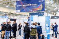 Auf der Messe SHK Essen stehen Menschen zusammen, diskutieren und tauschen Ideen aus. Im Hintergrund ist ein Banner mit der Aufschrift "STARTUP@SHK+E ESSEN" zu sehen. Stände präsentieren verschiedene Produkte und Innovationen.