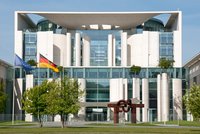 Das Bild zeigt die Fassade eines modernen Regierungsgebäudes mit großen Glasflächen und weißen Säulen. Vor dem Gebäude stehen Bäume und Skulpturen. Links wehen die Flaggen der Europäischen Union und Deutschlands. Der Himmel ist klar und blau.