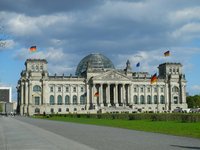 Das Bild zeigt das Reichstagsgebäude in Berlin, ein historisches und bedeutendes Gebäude der deutschen Politik. Es hat eine markante Glaskuppel und wird von mehreren Fahnen geschmückt. Im Vordergrund sieht man eine breite Promenade und Menschen, die sich auf dem Gelände bewegen.