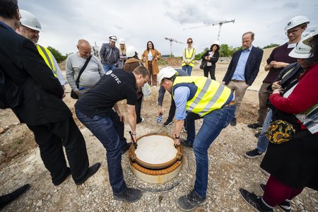 Zwei Personen heben gemeinsam eine runde Abdeckung auf einer Baustelle an, während eine Gruppe von Zuschauern in Sicherheitshelmen zusieht. Im Hintergrund sind Baukräne und eine unbebaute Landschaft zu sehen.