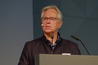 Ein älterer Mann mit grauen Haaren und Brille spricht konzentriert am Mikrofon. Er trägt einen blauen Pullover und eine dunkle Jacke. Im Hintergrund ist eine graue Wand zu sehen. Der Mann scheint Informationen oder Ideen zu präsentieren.