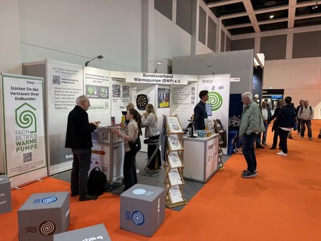 Messestand auf einer Veranstaltung mit einem Informationsstand des Bundesverbands Wärmepumpe (BWP) e.V. Besucher interagieren am Stand, der mit QR-Codes und Informationsmaterial ausgestattet ist. Der Boden ist orangefarben, und hinter dem Stand sind weitere Stände zu erkennen.