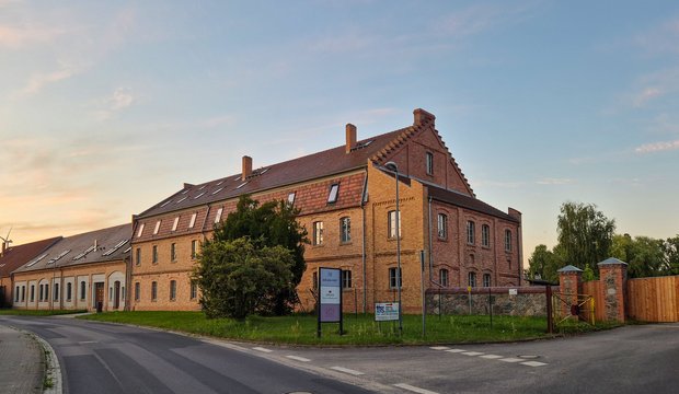 Das Bild zeigt ein ehemaliges, zweigeschössiges Backsteingebäude am Straßenrand, das gepflasterte Wege und Rasenflächen umgibt. Die Architektur weist eine Kombination aus traditionellen Elementen und modernen Fenstern auf. Im Hintergrund ist ein klarer Himmel zu sehen.