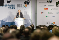 Eine Frau steht an einem Rednerspult und spricht vor einem Publikum bei der Neujahrsempfang-Veranstaltung des Bundesverbands Erneuerbare Energie (BEE) im Jahr 2019. Im Hintergrund sind Sponsorenlogos und eine stilisierte Grafik zu sehen.