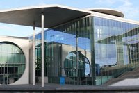 Das Bild zeigt ein modernes, architektonisch ansprechendes Gebäude mit großen Glasflächen und einer charakteristischen Dachform. Es sind auch Säulen sichtbar, die das Dach stützen, sowie eine Treppe im Vordergrund. Das Gebäude reflektiert das Licht und die Umgebung.
