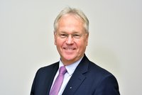 Porträt eines lächelnden Mannes mit kurzen, grauen Haaren, der eine Brille trägt. Er trägt einen dunkelblauen Anzug und ein lila gemustertes Krawatte. Der Hintergrund ist hell und neutral gehalten.