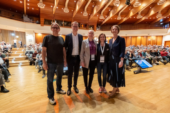 Fünf Personen stehen auf einer Bühne in einem großen Saal, der mit Zuschauern gefüllt ist. Sie lächeln freundlich in die Kamera. Die Bühne ist mit modernen Technologien ausgestattet und hat helle Holzelemente, während die Zuschauersitze im Hintergrund zu sehen sind.