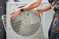 Ein Handwerker entfernt ein Gitter von einer Klimaanlage. Die Person trägt eine Arbeitskleidung und hält das Gitter mit beiden Händen. Im Hintergrund ist die Klimaanlage sichtbar.
