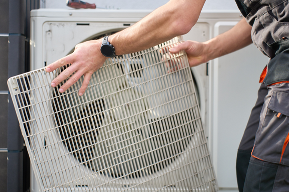 Ein Handwerker entfernt ein Gitter von einer Klimaanlage. Die Person trägt eine Arbeitskleidung und hält das Gitter mit beiden Händen. Im Hintergrund ist die Klimaanlage sichtbar.