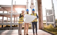 Drei Fachleute in Sicherheitswesten und Helmen stehen auf einer Baustelle und schauen sich einen Bauplan an. Im Hintergrund sind unvollendete Betonstrukturen sichtbar, und die Sonne scheint. Die Personen diskutieren konzentriert über das Projekt.