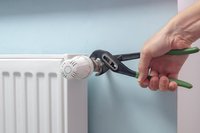 Eine Hand hält eine Gripzange, die an einem Heizungsventil ansetzt. Im Hintergrund ist ein hellblauer Wandbereich und ein weißer Heizkörper sichtbar. Die Zange wird verwendet, um Anpassungen am Thermostat vorzunehmen.