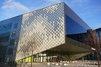 Ein modernes Gebäude mit einer auffälligen, diamantförmigen Fassade reflektiert das Licht. Der Eingang ist verdeckt, mit großen Fenstern und einem grünen Bereich. Vor dem Gebäude stehen mehrere Fahrräder, während eine schmale Baumgruppe auf der linken Seite sichtbar ist.