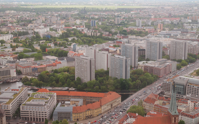 Luftaufnahme einer Stadtlandschaft mit modernen Wohngebäuden und viel grüner Vegetation. Im Hintergrund sind weitere Gebäude und eine weitläufige Landschaft zu sehen. Straßen mit Fahrzeugen verlaufen zwischen den Gebäuden, und die städtische Struktur wird von Parks und Bäumen unterbrochen.