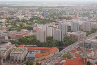 Luftaufnahme einer Stadtlandschaft mit modernen Wohngebäuden und viel grüner Vegetation. Im Hintergrund sind weitere Gebäude und eine weitläufige Landschaft zu sehen. Straßen mit Fahrzeugen verlaufen zwischen den Gebäuden, und die städtische Struktur wird von Parks und Bäumen unterbrochen.