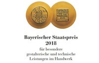 Zwei Medaillen mit Text: Eine zeigt ein symbolisches Design, die andere den Schriftzug "Bayerischer Staatspreis für das Handwerk 2018". Der Preis wird für besondere gestalterische und technische Leistungen im Handwerk vergeben.