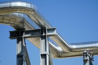 Eine Gruppe von großen, glänzenden Metallrohren verläuft über eine graue tragende Struktur. Die Rohre winden sich unter einem klaren blauen Himmel und zeigen eine moderne industrielle Konstruktion.