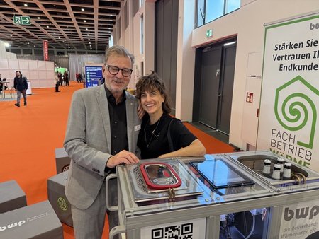 Zwei Personen stehen an einem Infostand auf einer Messe. Der Mann trägt einen grauen Anzug und eine Brille, die Frau hat kurze, lockige Haare und trägt ein schwarzes Oberteil. Auf dem Stand sind Geräte und Informationstafeln zu sehen. Im Hintergrund sind Messebesucher und orangefarbener Boden.