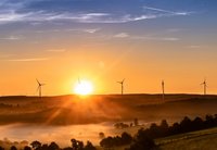 Sonnenaufgang über einer hügeligen Landschaft, umgeben von mehreren Windkraftanlagen. Der Himmel zeigt sanfte Farbverläufe von Orange bis Blau. Sanfte Nebelschwaden liegen über dem Land, während die Sonne langsam aufgeht und die Umgebung in warmes Licht taucht.