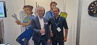 Zwei Männer stehen lächelnd vor einem Hintergrund mit Zeichnungen von Handwerkern. Der Mann links zeigt ein Daumen hoch, während der andere freundlich schaut. Im Hintergrund sind Werkzeuge und ein Schild mit der Aufschrift „GET FIT“ zu sehen.