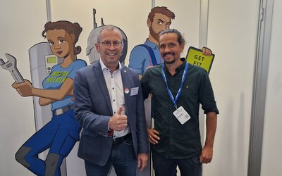 Zwei Männer stehen lächelnd vor einem Hintergrund mit Zeichnungen von Handwerkern. Der Mann links zeigt ein Daumen hoch, während der andere freundlich schaut. Im Hintergrund sind Werkzeuge und ein Schild mit der Aufschrift „GET FIT“ zu sehen.