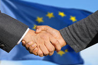 Zwei Hände schütteln sich vor einem unscharfen Hintergrund mit der Flagge der Europäischen Union. Die Geste symbolisiert Zusammenarbeit und Einigung.