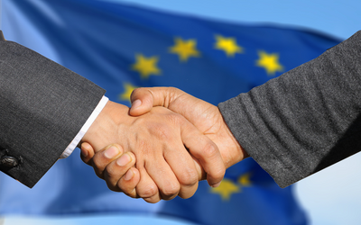 Zwei Hände schütteln sich vor einem unscharfen Hintergrund mit der Flagge der Europäischen Union. Die Geste symbolisiert Zusammenarbeit und Einigung.
