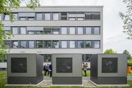 Drei große graue Maschinen stehen im Vordergrund vor einem modernen, mehrstöckigen Gebäude mit vielen Fenstern. Im Hintergrund sind mehrere Personen zu sehen, die sich unterhalten. Bäume und Gras sind ebenfalls sichtbar.