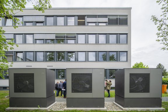 Drei große graue Maschinen stehen im Vordergrund vor einem modernen, mehrstöckigen Gebäude mit vielen Fenstern. Im Hintergrund sind mehrere Personen zu sehen, die sich unterhalten. Bäume und Gras sind ebenfalls sichtbar.