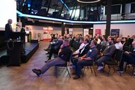 Eine Präsentation findet in einem modernen Raum statt. Ein Redner steht an einem Pult und spricht zu einem Publikum von Männern und Frauen, die in Reihen sitzen. Das Publikum hört aufmerksam zu, einige machen sich Notizen.