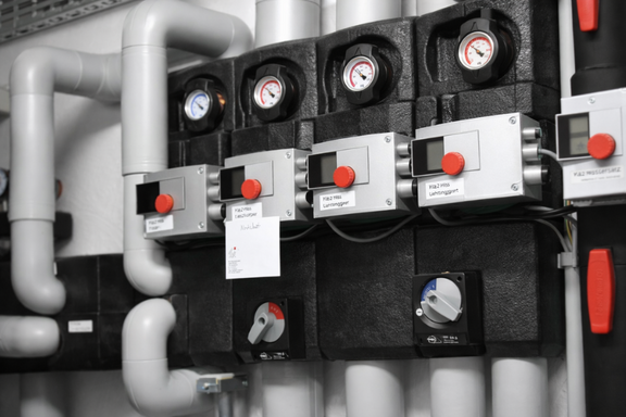 Bildbeschreibung: Ein technisches Steuerungs- und Messsystem mit mehreren schwarzen Steuergeräten und Manometern. Verschiedene Rohre, sowohl in grau als auch in weiß, sind sichtbar, die an die Geräte angeschlossen sind. Es gibt rot markierte Knöpfe und digitale Anzeigen zur Überwachung.