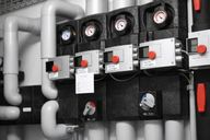 Bildbeschreibung: Ein technisches Steuerungs- und Messsystem mit mehreren schwarzen Steuergeräten und Manometern. Verschiedene Rohre, sowohl in grau als auch in weiß, sind sichtbar, die an die Geräte angeschlossen sind. Es gibt rot markierte Knöpfe und digitale Anzeigen zur Überwachung.