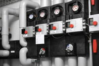 Bildbeschreibung: Ein technisches Steuerungs- und Messsystem mit mehreren schwarzen Steuergeräten und Manometern. Verschiedene Rohre, sowohl in grau als auch in weiß, sind sichtbar, die an die Geräte angeschlossen sind. Es gibt rot markierte Knöpfe und digitale Anzeigen zur Überwachung.