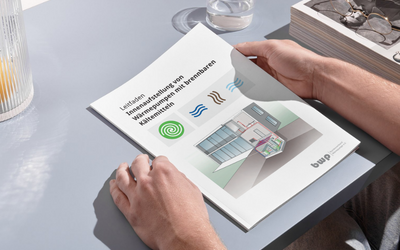 Eine Person hält ein Dokument mit dem Titel „Leitfaden Immersionsdüsen von Wärmepumpen mit Innenmaschinen“ in den Händen. Das Blatt zeigt eine Illustration mit Symbolen und einem grafischen Diagramm, das einen Raum und Zubehör beschreibt. Der Hintergrund enthält einen Tisch mit einer Brille und einem Glas Wasser.