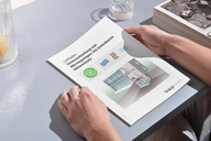 Eine Person hält ein Dokument mit dem Titel „Leitfaden Immersionsdüsen von Wärmepumpen mit Innenmaschinen“ in den Händen. Das Blatt zeigt eine Illustration mit Symbolen und einem grafischen Diagramm, das einen Raum und Zubehör beschreibt. Der Hintergrund enthält einen Tisch mit einer Brille und einem Glas Wasser.