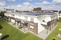 Eine moderne Wohnanlage mit mehreren Wohnungen, die über Balkone verfügen. Auf dem Dach sind Solarpanels installiert. Im Vordergrund erstreckt sich eine grüne Rasenfläche. Der Himmel ist teilweise bewölkt. Die Gebäude sind in hellen und dunklen Farbtönen gehalten.