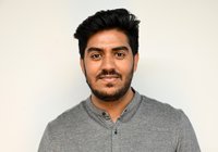 Ein junger Mann mit schwarzem Haar und einem kurzen Bart lächelt in die Kamera. Er trägt ein graues Hemd. Der Hintergrund ist neutral und hell.