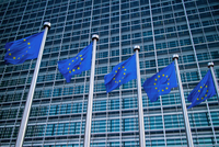 Fünf Flaggen der Europäischen Union wehen vor einem modernen Gebäude mit Glasfassade. Die Flaggen zeigen den blauen Hintergrund mit gelben Sternen, charakteristisch für die EU.