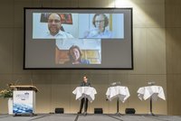 Eine Moderatorin steht auf der Bühne vor einem großen Bildschirm, auf dem drei Personen in Videoanrufen zu sehen sind. Die Veranstaltung scheint eine Diskussion oder Präsentation zu sein, die Technologie zur Verbindung von Teilnehmern nutzt. Zwei der Personen haben Headsets. Tische mit Wasserflaschen sind im Vordergrund.