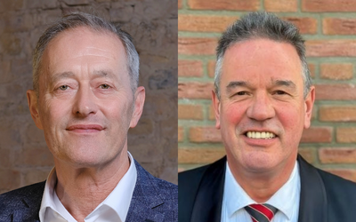 Das Bild zeigt zwei Männer, jeweils zur Hälfte abgebildet. Der Mann links trägt ein helles Hemd und einen dunkelblauen Blazer, während der Mann rechts einen Anzug mit Krawatte trägt. Beide lächeln direkt in die Kamera.