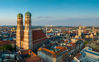 Blick auf die Münchener Altstadt mit den markanten Zwillingstürmen der Frauenkirche im Vordergrund. Die umliegenden Dächer und historischen Gebäude sind in warmes Licht getaucht, während der Himmel klar und blau ist.