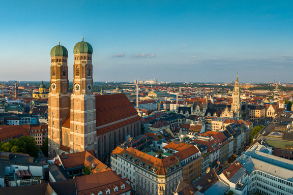 Blick auf die Münchener Altstadt mit den markanten Zwillingstürmen der Frauenkirche im Vordergrund. Die umliegenden Dächer und historischen Gebäude sind in warmes Licht getaucht, während der Himmel klar und blau ist.