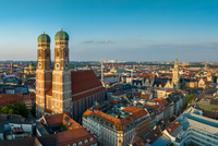 Blick auf die Münchener Altstadt mit den markanten Zwillingstürmen der Frauenkirche im Vordergrund. Die umliegenden Dächer und historischen Gebäude sind in warmes Licht getaucht, während der Himmel klar und blau ist.
