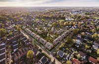 Luftaufnahme eines Wohngebiets mit modernen Reihenhäusern, umgeben von Bäumen und Landschaft. Die Häuser sind unterschiedlich angeordnet und verfügen über Gärten. Im Hintergrund sind weitere Wohnhäuser und die natürliche Umgebung sichtbar. Das Bild vermittelt ein Gefühl von Ruhe und Gemeinschaft.