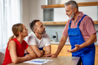 Drei Personen sitzen an einem Tisch in einer modernen Küche. Ein älterer Mann in blauer Arbeitskleidung spricht gestikulierend mit einem Paar, das aufmerksam zuhört. Auf dem Tisch liegen Unterlagen und Schreibutensilien. Die Atmosphäre vermittelt ein Gespräch über Renovierungsarbeiten oder Dienstleistungen.